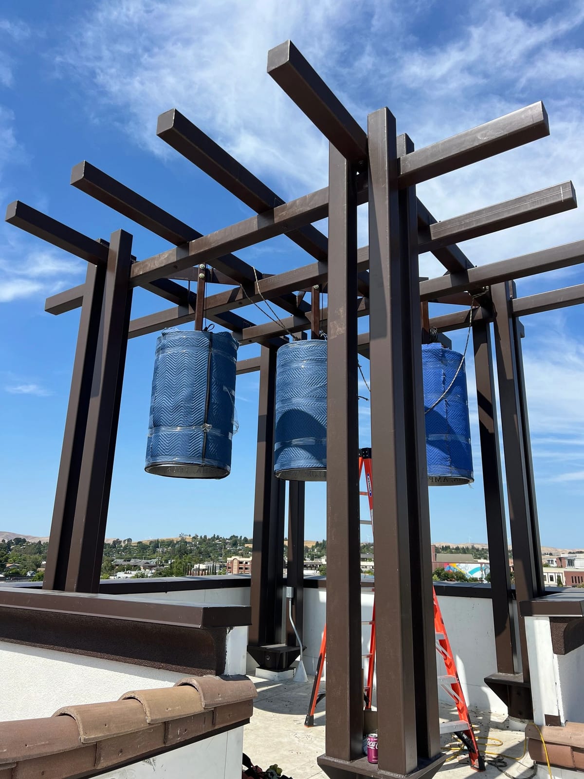 Rooftop bell tower trellis of dark bronze painted steel posts and crossing beams, with three bells hanging wrapped in blue blankets