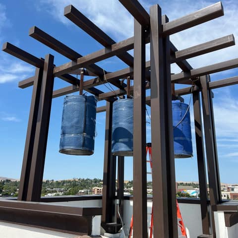 Rooftop bell tower trellis of dark bronze painted steel posts and crossing beams, with three bells hanging wrapped in blue blankets
