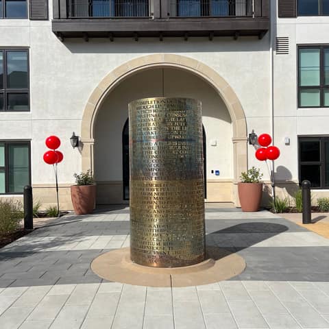 Large brass-finish cylinder with laser-cut historical text, standing in the entry plaza of an apartment building in Livermore.