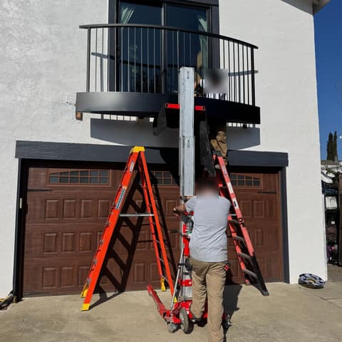 Curved black steel balcony with round vertical pickets being installed above a garage using a material lift.