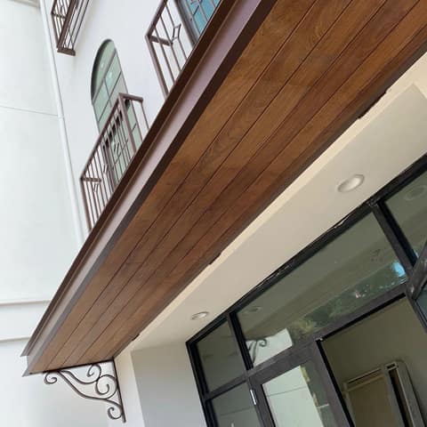 Decorative entry awning with a dark bronze painted steel frame, wood plank underside and a scrolled iron bracket, below matching bronze window railings