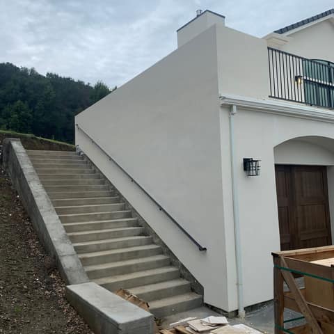 Black steel wall-mounted handrail along a long exterior concrete stair beside a new home.