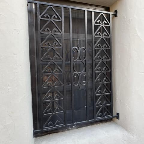 Black wrought iron window guard with hand-formed triangular scroll patterns and a center scroll medallion.