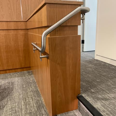 Brushed stainless steel pipe handrail on wall brackets beside a wood-paneled stage step.