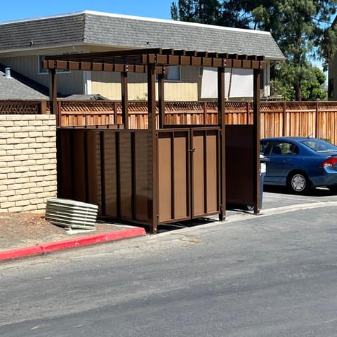 Brown steel trash enclosure with solid panel walls, double doors and a slatted trellis top in an apartment parking lot.