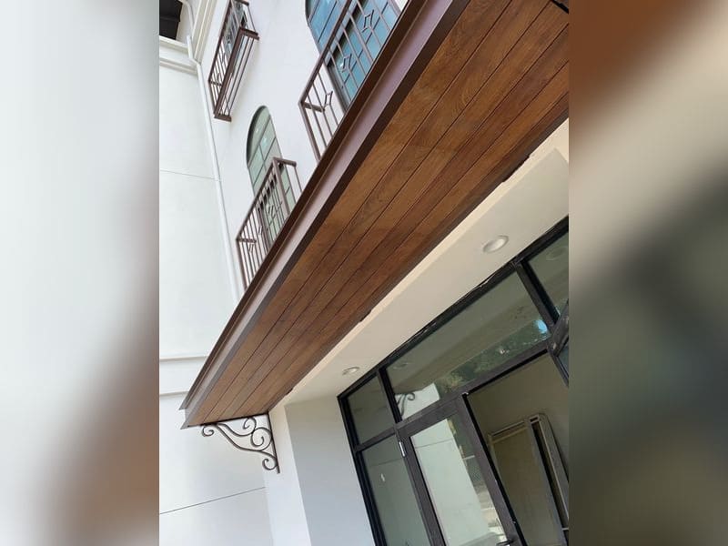 Decorative entry awning with a dark bronze painted steel frame, wood plank underside and a scrolled iron bracket, below matching bronze window railings