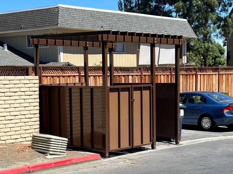 Brown steel trash enclosure with solid panel walls, double doors and a slatted trellis top in an apartment parking lot.
