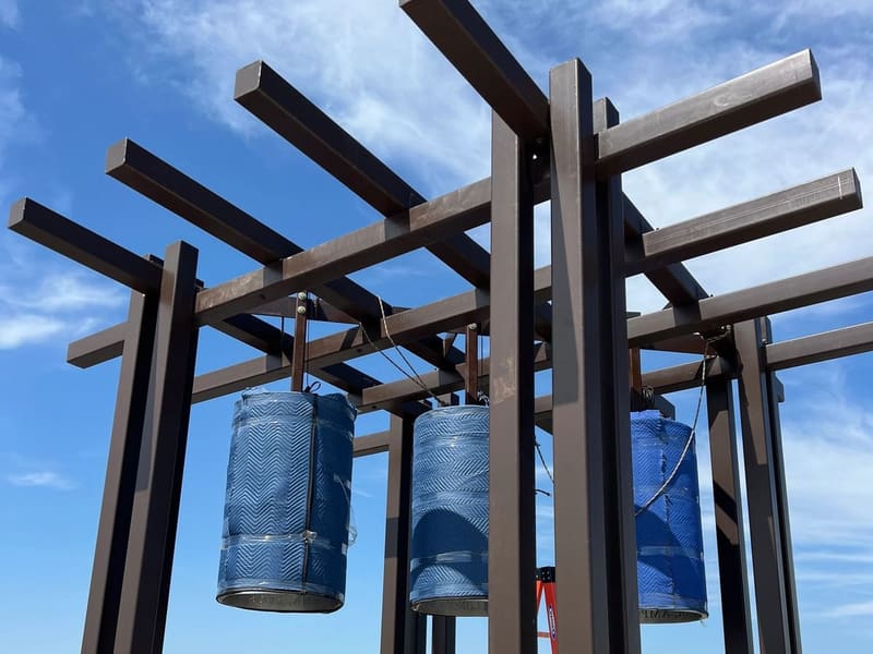 Rooftop bell tower trellis of dark bronze painted steel posts and crossing beams, with three bells hanging wrapped in blue blankets